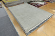 A large rectangular area rug in a light gray and white ribbed pattern with a dark gray patterned border, displayed on a light wood floor in a showroom with other rugs visible.