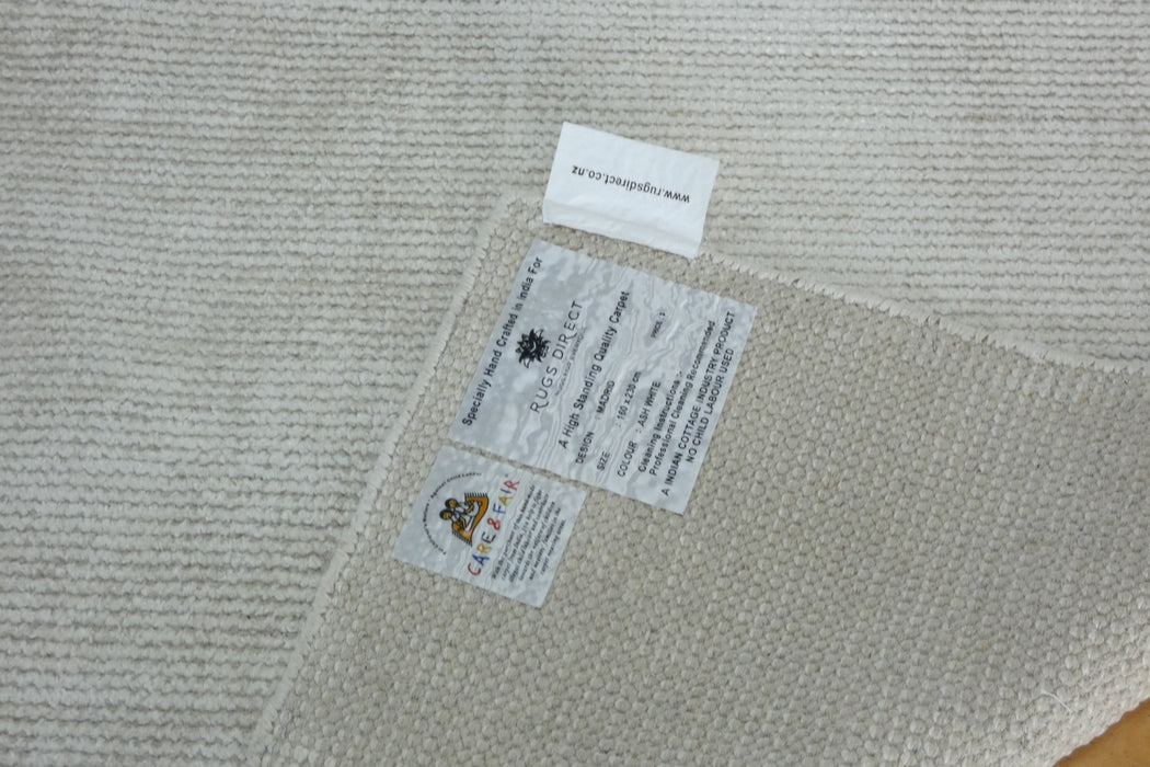 Close-up of a woven off-white rug corner showing two sewn-in labels, including a circular logo that appears to read 'The Rug Club' and a small white tag above it.
