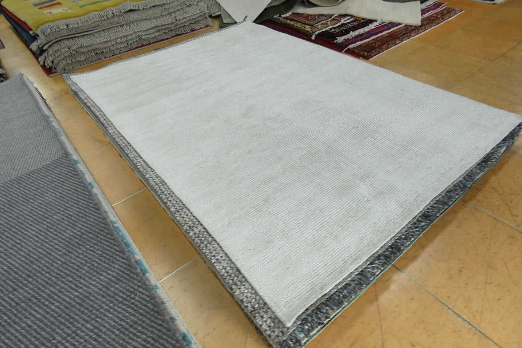 A large rectangular area rug in a light gray to off-white tone with a textured, ribbed surface and a dark gray patterned border, displayed on a tiled floor in what appears to be a showroom or warehouse.