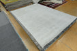A large rectangular area rug in a light gray to off-white tone with a textured, ribbed surface and a dark gray patterned border, displayed on a tiled floor in what appears to be a showroom or warehouse.