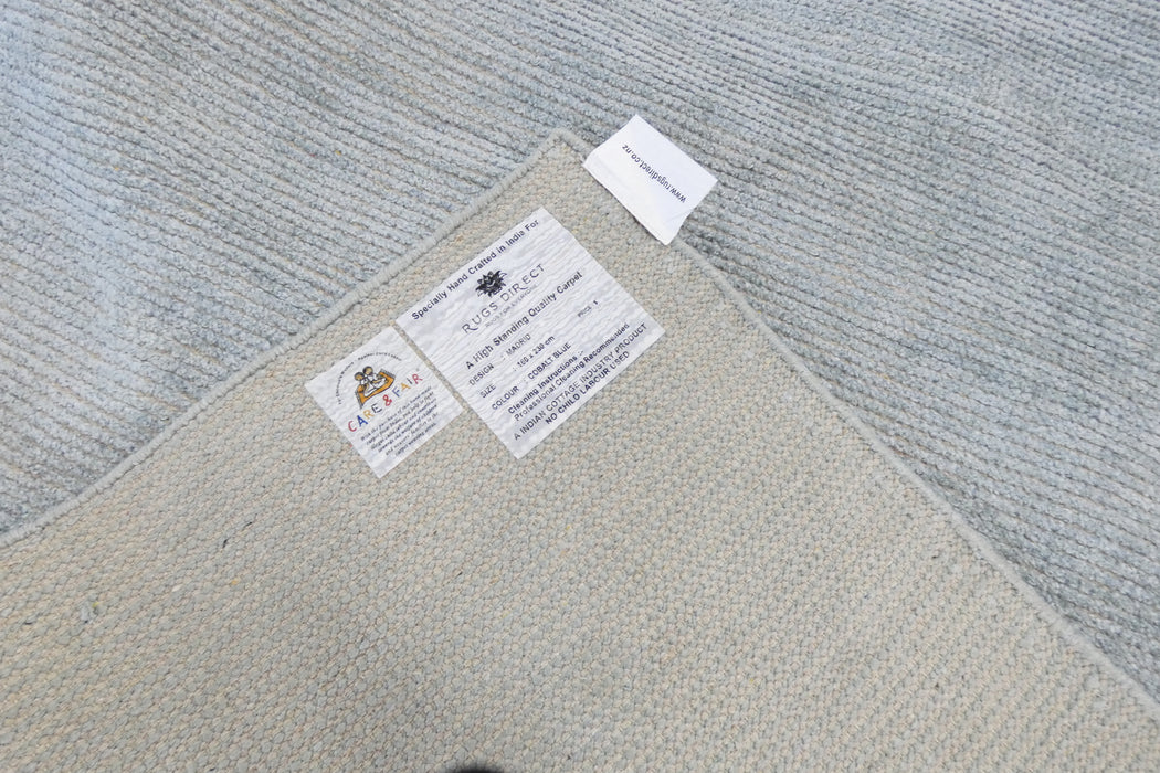 Close-up of the corner of a woven area rug showing the backing, product labels, and the adjacent light gray textured rug surface.