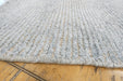 A close-up of a textured, chunky knit rug in a light gray and white heathered pattern, laid out on a light-colored wooden floor.