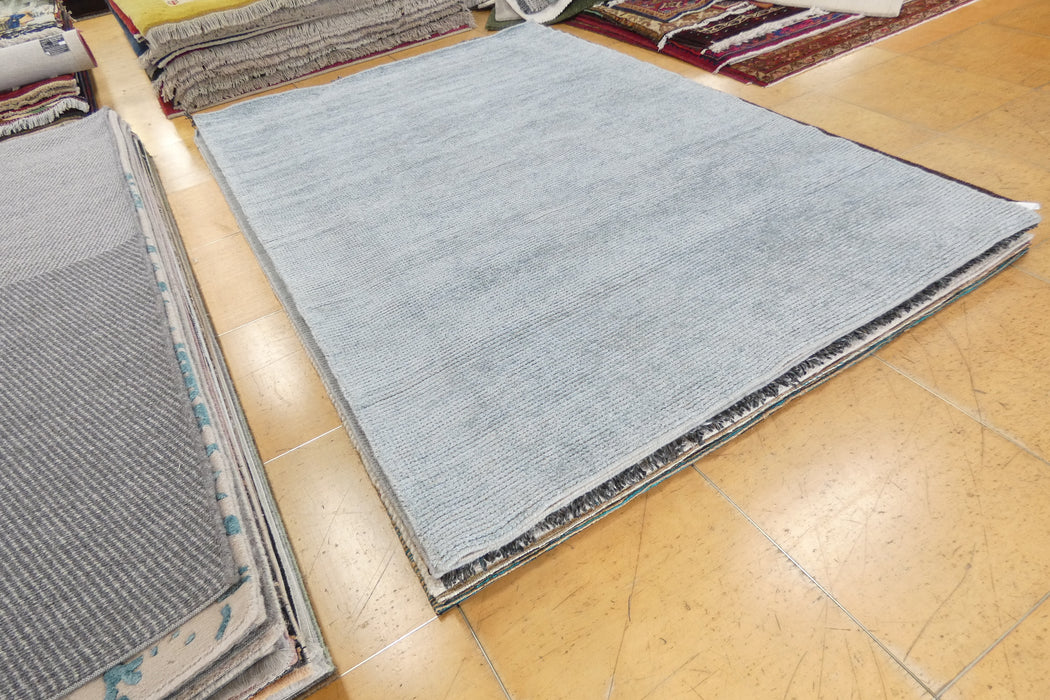 A large light blue-gray rectangular rug laid out on a tan tiled floor, with stacks of other rugs visible around it in a showroom.