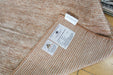 A close-up of a textured woven rug or carpet in a warm beige and brown houndstooth pattern, with two white product labels attached near the folded corner.