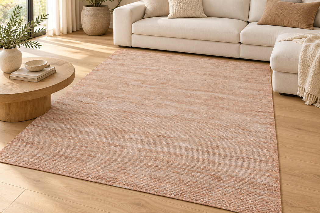 A rectangular area rug in a warm dusty rose or blush pink color, styled in a bright, modern living room with a light wood floor, a cream sofa, and a round wooden coffee table.