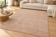 A rectangular area rug in a warm dusty rose or blush pink color, styled in a bright, modern living room with a light wood floor, a cream sofa, and a round wooden coffee table.