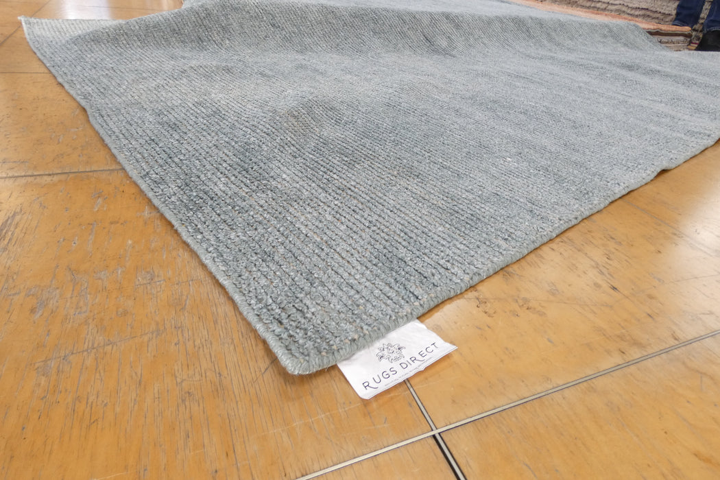A light gray textured rug laid out on a polished wooden floor, with a white fabric tag reading 'RUGS DIRECT' visible near one corner.