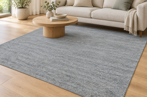 A large gray textured area rug laid out on a light wood floor in a modern living room setting with a beige sofa, round wooden coffee table, and neutral decor.