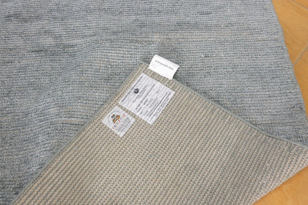 A close-up of a gray textured rug with a folded corner showing a beige underside and two product labels, placed on a light wood floor.