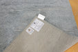 A close-up of a gray textured rug with a folded corner showing a beige underside and two product labels, placed on a light wood floor.