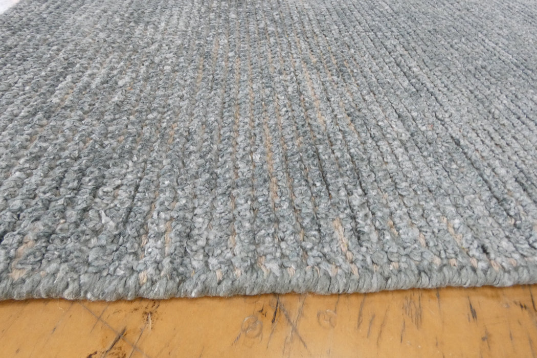A close-up of a gray textured rug with a chunky, braided or boucle-like surface, shown resting on a light brown wooden floor.