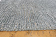 A close-up of a gray textured rug with a chunky, braided or boucle-like surface, shown resting on a light brown wooden floor.