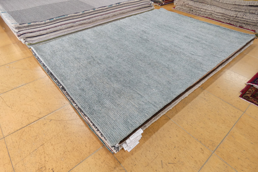 A large rectangular area rug in a muted blue-gray color, shown partially unrolled on a tan tiled floor with other stacked rugs visible in the background.