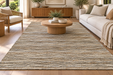 A large rectangular area rug with a textured, heathered pattern in beige, brown, and cream tones, styled in a bright, modern living room with a wooden coffee table, white sofa, and wicker chair.