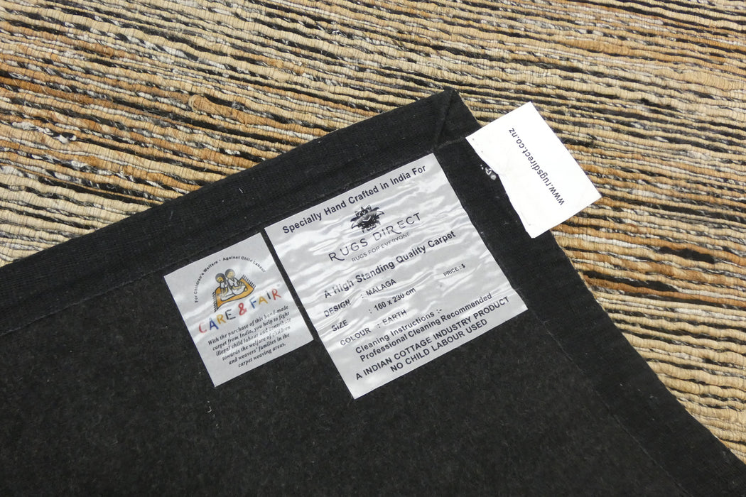 A close-up of a black carpet sample with two product labels attached, placed on a textured tan and brown woven rug. The labels show the brand 'Ridge-Rift' and the manufacturer 'A&F & F'.