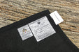 A close-up of a black carpet sample with two product labels attached, placed on a textured tan and brown woven rug. The labels show the brand 'Ridge-Rift' and the manufacturer 'A&F & F'.