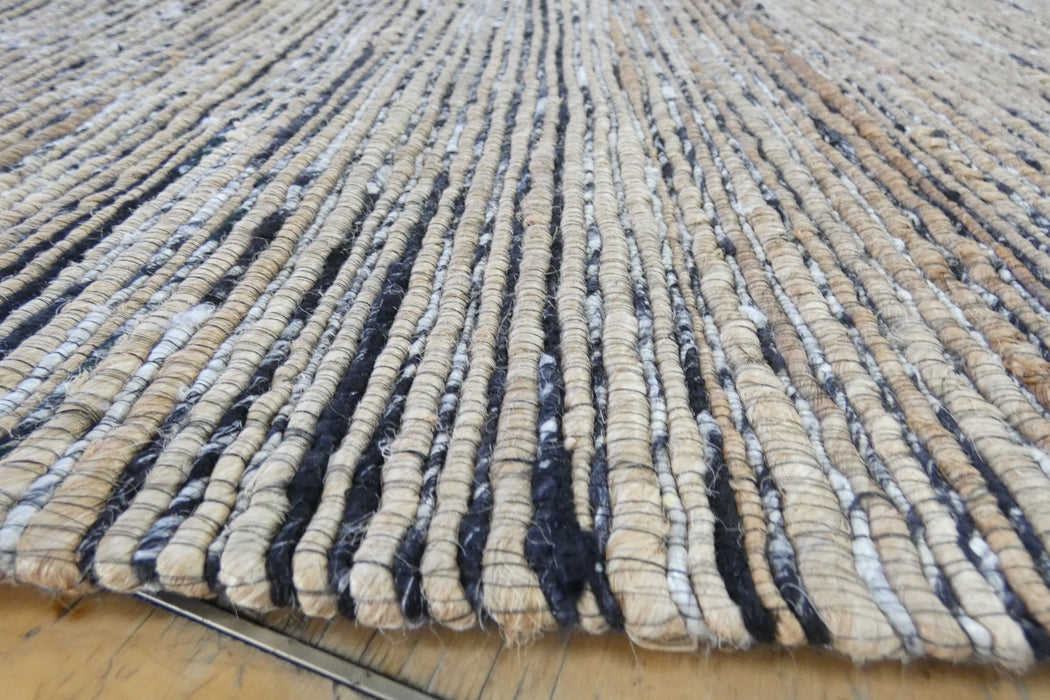 A close-up of a woven rug with vertical textured stripes in beige, brown, black, and white, shown on a light wood floor.