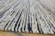A close-up of a woven rug with vertical textured stripes in beige, brown, black, and white, shown on a light wood floor.