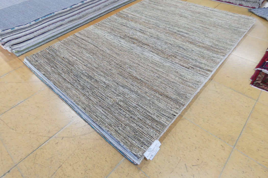 A rectangular woven area rug with a textured, heathered pattern in shades of beige, gray, and brown, displayed on a light tan tiled floor next to other rugs.