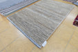 A rectangular woven area rug with a textured, heathered pattern in shades of beige, gray, and brown, displayed on a light tan tiled floor next to other rugs.
