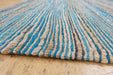 A close-up of a woven rug with vertical textured stripes in turquoise blue, natural beige, and dark brown, shown on a light wood floor.