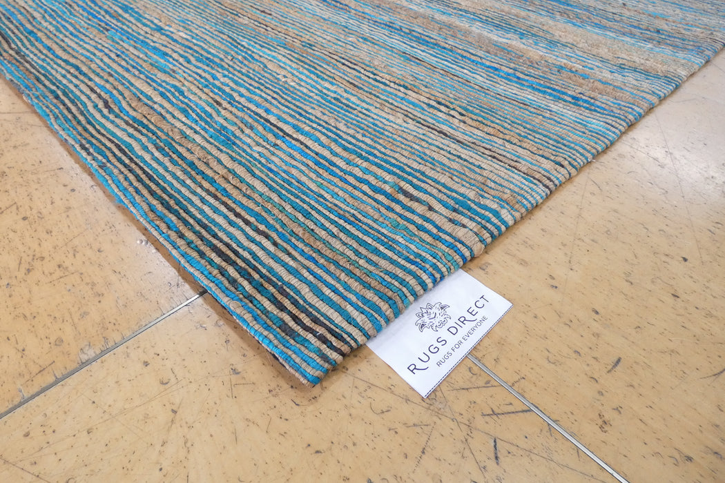 A close-up of a woven area rug corner on a light wood floor, showing a blue, beige, and brown striped pattern with a white 'RUGS DIRECT' tag attached.