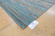 A close-up of a woven area rug corner on a light wood floor, showing a blue, beige, and brown striped pattern with a white 'RUGS DIRECT' tag attached.