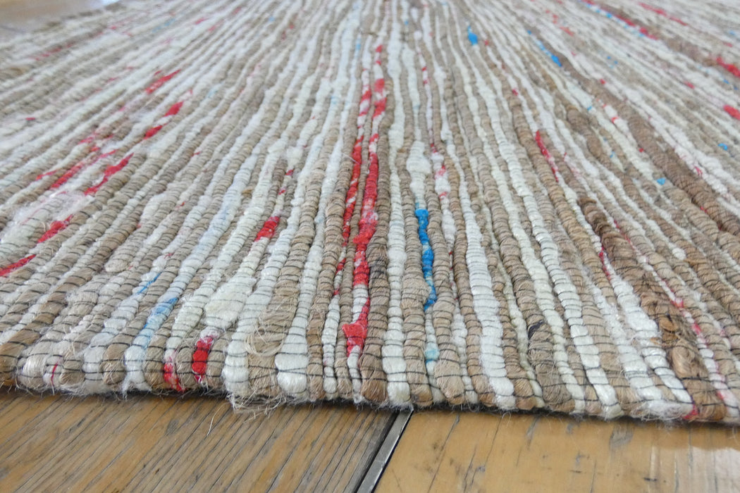 A close-up of a woven shag rug made from natural jute fibers, featuring vertical stripes in beige, off-white, and small accents of red and blue, laid on a wooden floor.