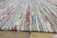 A close-up of a woven shag rug made from natural jute fibers, featuring vertical stripes in beige, off-white, and small accents of red and blue, laid on a wooden floor.