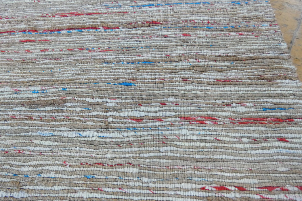 A close-up of a woven textile or rug with horizontal natural-fiber stripes in beige, white, and small scattered red and blue accents.