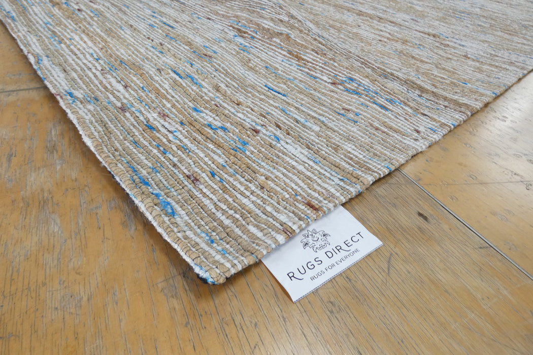 Close-up of a woven rug corner on a light wood floor, showing a white tag that reads RUGS DIRECT. The rug has a natural beige and off-white jute base with subtle blue and brown flecks.