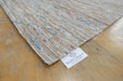 Close-up of a woven rug corner on a light wood floor, showing a white tag that reads RUGS DIRECT. The rug has a natural beige and off-white jute base with subtle blue and brown flecks.