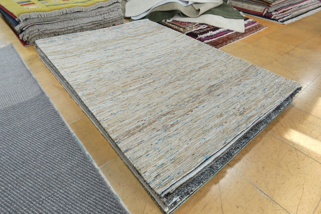 A rectangular woven area rug in a light beige and gray tone with subtle blue flecks, displayed on a wooden floor alongside other rugs.