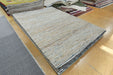 A rectangular woven area rug in a light beige and gray tone with subtle blue flecks, displayed on a wooden floor alongside other rugs.