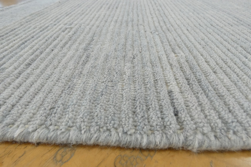 A close-up view of a light gray ribbed rug with a frayed edge, laid on a yellowish wooden floor.