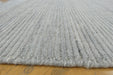 A close-up view of a light gray ribbed rug with a frayed edge, laid on a yellowish wooden floor.