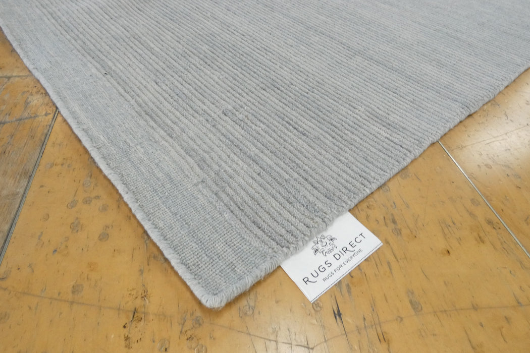 A close-up of a light gray ribbed area rug laid on a worn yellow-brown wooden floor, with a white 'RUGS DIRECT' brand tag visible at the corner.