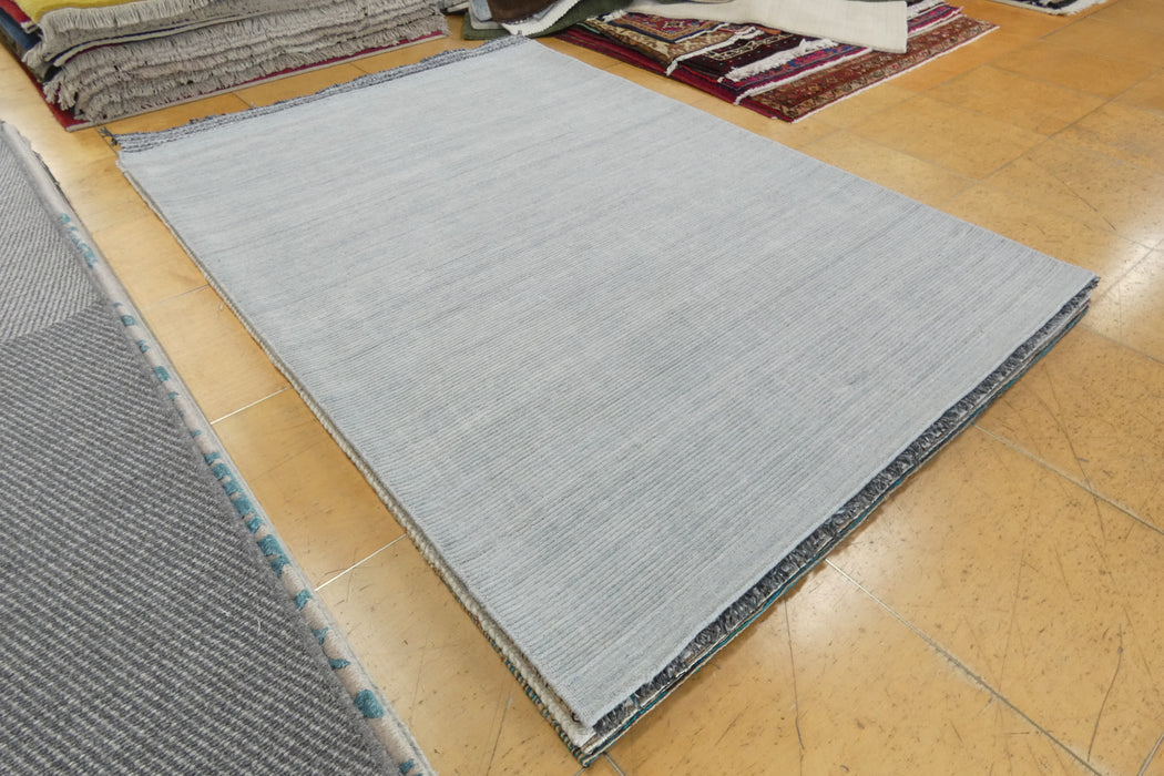 A light gray rectangular area rug laid out on a tan tiled floor, with other rugs stacked in the background.