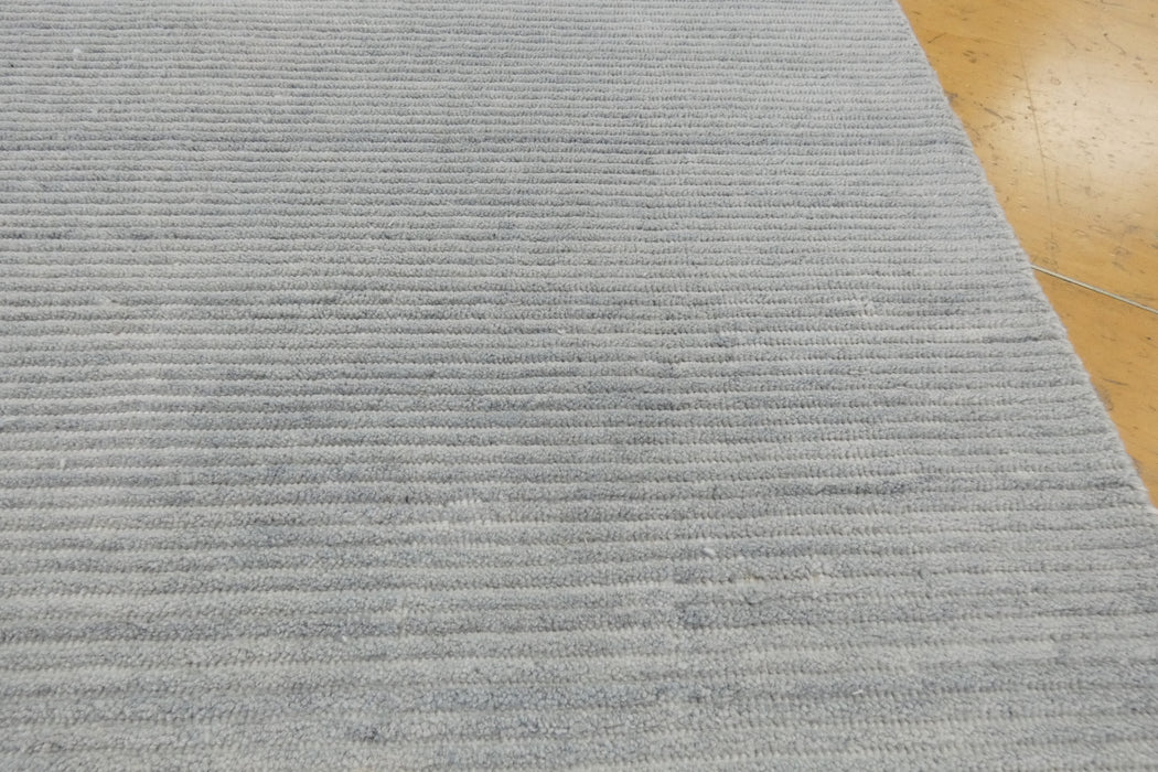 A close-up of a light gray textured rug with horizontal ribbed stripes, shown with a portion of a light brown wooden floor visible in the top right corner.