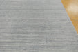 A close-up of a light gray textured rug with horizontal ribbed stripes, shown with a portion of a light brown wooden floor visible in the top right corner.
