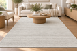 A large rectangular area rug in a light gray and white textured pattern, shown in a bright, modern living room with a wooden coffee table, beige sofa, and wood flooring.