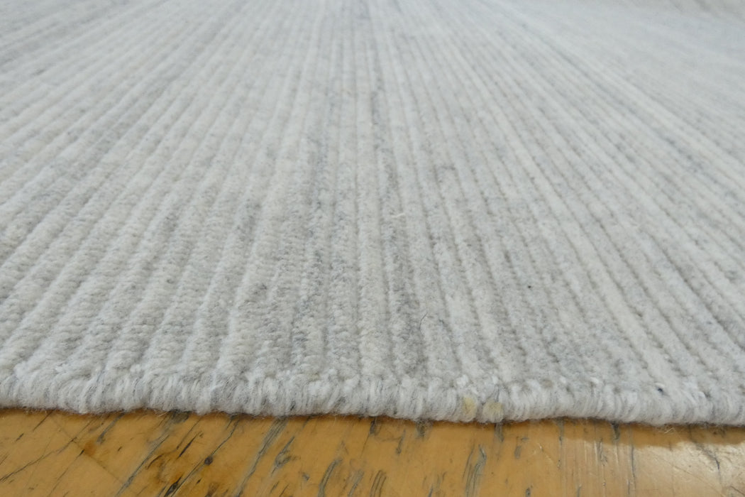 Close-up of a light gray ribbed wool or felt rug with a fringed edge, shown resting on a yellowish wooden floor.