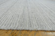 Close-up of a light gray ribbed wool or felt rug with a fringed edge, shown resting on a yellowish wooden floor.