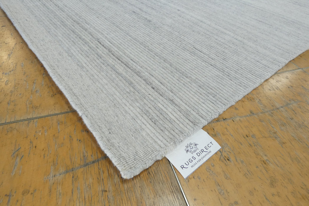 A close-up of a light gray textured rug corner with a visible white 'RUGS DIRECT' label, placed on a worn wooden floor.