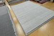 A large light gray rectangular area rug with a subtle horizontal ribbed texture, displayed on a tiled showroom floor alongside other rugs.