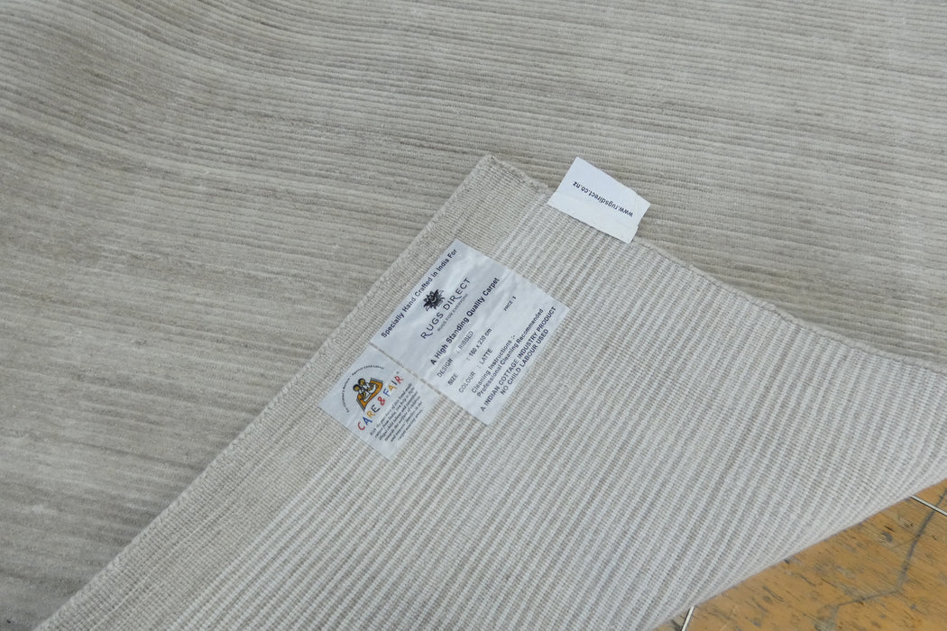 A close-up of a light beige textured area rug with a ribbed pattern, showing product labels on one corner and a small white tag, with a small portion of a wooden floor visible in the bottom right corner.