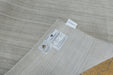 A close-up of a light beige textured area rug with a ribbed pattern, showing product labels on one corner and a small white tag, with a small portion of a wooden floor visible in the bottom right corner.