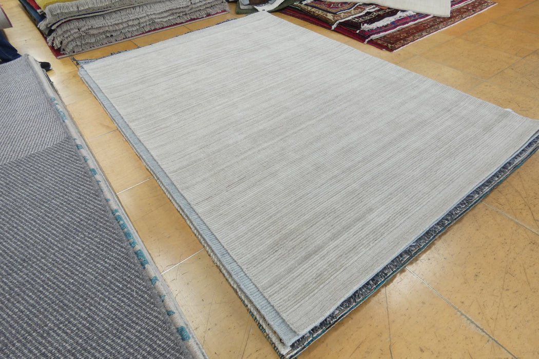 A large rectangular area rug with a light gray and white textured pattern, displayed on a tan tiled floor alongside other rugs.
