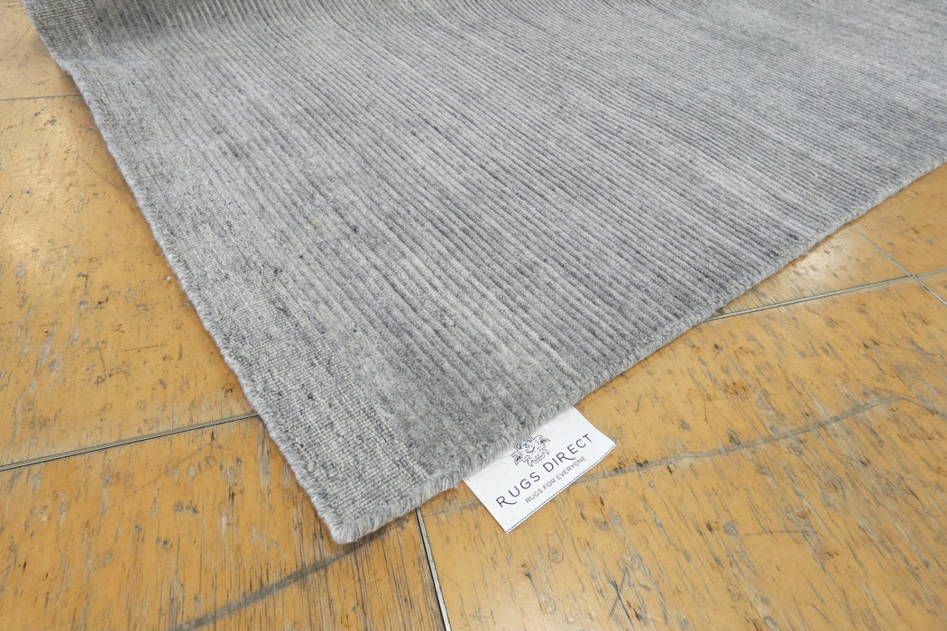 A close-up of a gray textured area rug laid on a light brown wooden floor, with a white Rug Direct brand tag visible at the corner.