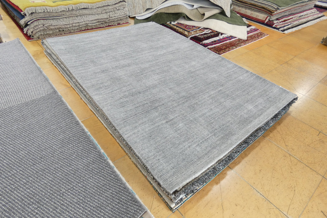 A rectangular light gray rug sample or finished rug shown on a wood floor, with stacks of folded rugs in the background.